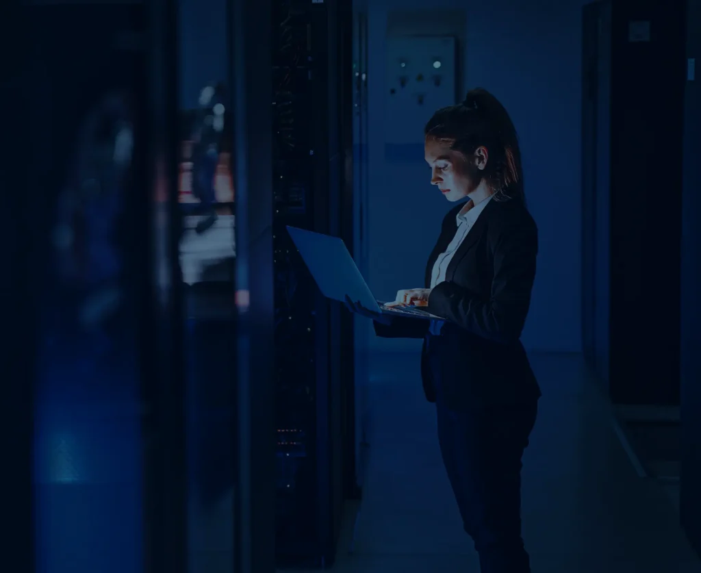 Businesswoman working on laptop in server room, illustrating IT support and cybersecurity solutions.