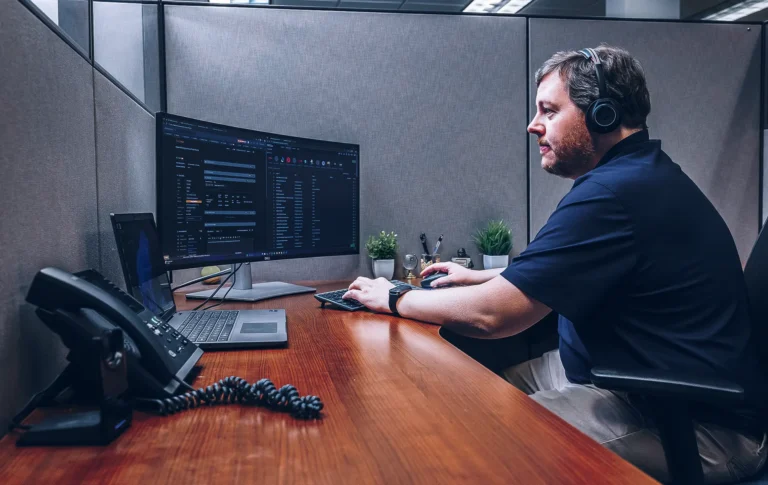 IT support technician at desk with headphones, monitoring computer screens and phone system.